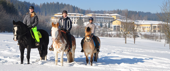 Winterlich zugeschneite Landschaft mit dem Hotel Aldiana Club Ampflwang in Österreich. Im Vordergrund stehen 3 Reiterinnen.