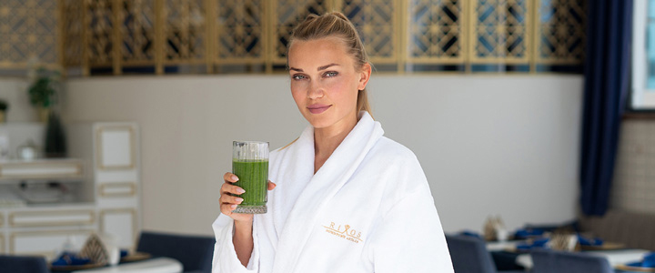 Frau steht im Bademantel in einem Café, hält einen grünen Smoothie in der Hand und schaut in die Kamera.